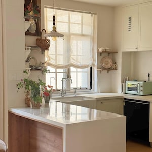 May include: A light and airy kitchen featuring a white countertop island with wooden detailing. A window with a cream-colored blind is above the sink. A mint green toaster sits on the counter. Decorative shelves and pendant lights enhance the space's charm.