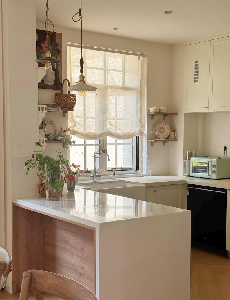May include: A light and airy kitchen featuring a white countertop island with wooden detailing. A window with a cream-colored blind is above the sink. A mint green toaster sits on the counter. Decorative shelves and pendant lights enhance the space's charm.