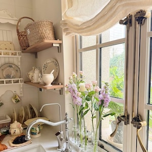 May include: A kitchen interior with a white sink and chrome tap, displaying shelves with decorative plates and a wicker basket. Vases of pink and purple flowers sit on the windowsill, next to a cream-coloured curtain.