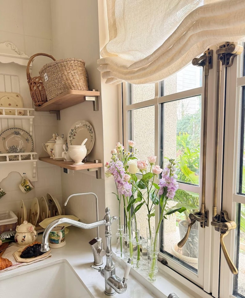 May include: A kitchen interior with a white sink and chrome tap, displaying shelves with decorative plates and a wicker basket. Vases of pink and purple flowers sit on the windowsill, next to a cream-coloured curtain.
