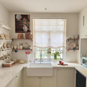 May include: A light and airy kitchen featuring white cabinetry, a farmhouse sink, and a window with a sheer blind. Shelves display decorative plates, cups, and a floral painting, creating a welcoming feel. Two apples rest on a wooden chopping board on the work surface.