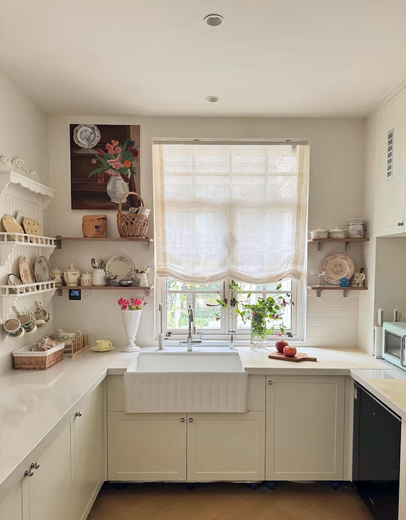 May include: A light and airy kitchen featuring white cabinetry, a farmhouse sink, and a window with a sheer blind. Shelves display decorative plates, cups, and a floral painting, creating a welcoming feel. Two apples rest on a wooden chopping board on the work surface.