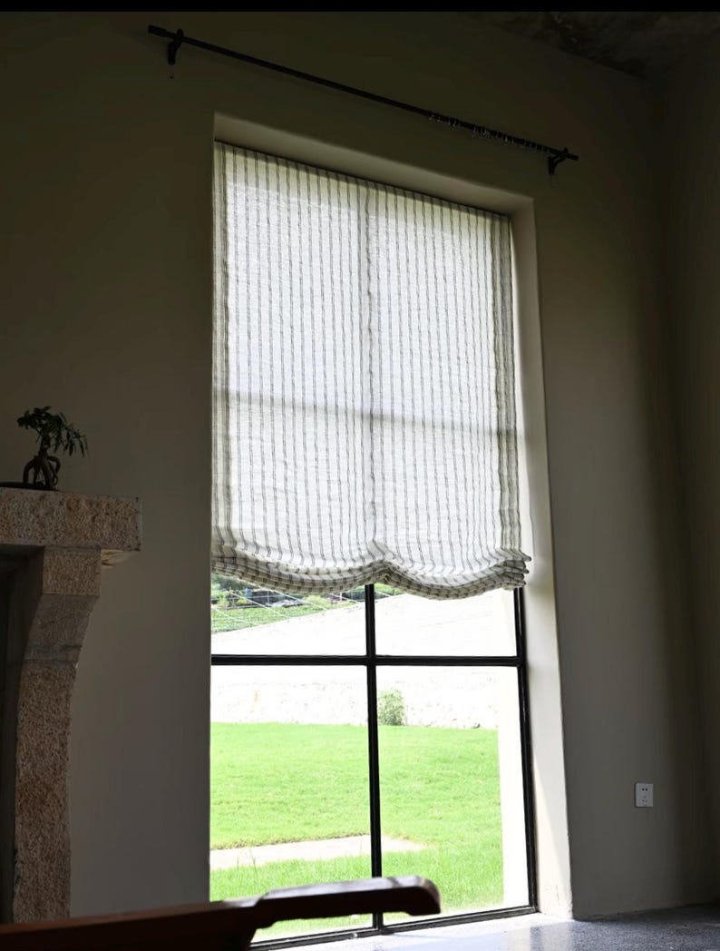 May include: A large window with a black grid frame and a striped, light-colored curtain. The curtain is pulled up, revealing a view of green grass outside. A black rod hangs above the window.