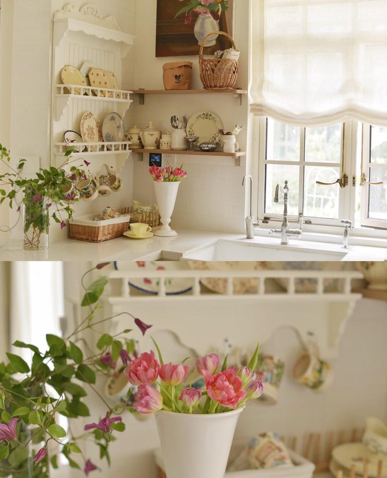 May include: A light and airy kitchen with white shelves displaying decorative plates and cups. A white vase holds pink tulips, and a window with a white curtain is in the background. A basket and a painting are also visible.
