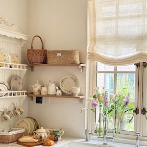 May include: A light and airy kitchen with white walls and shelves, featuring floral plates, teacups, and a teapot. A window with a linen blind overlooks a garden, with fresh flowers in vases. Baskets and bread complete the scene.