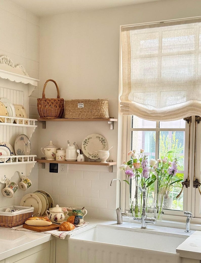 May include: A light and airy kitchen with white walls and shelves, featuring floral plates, teacups, and a teapot. A window with a linen blind overlooks a garden, with fresh flowers in vases. Baskets and bread complete the scene.
