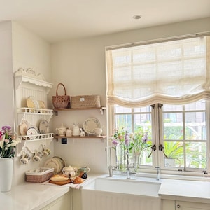 May include: A light and airy kitchen with white cabinets, a farmhouse sink, and a window with a linen blind. Decorative shelves display plates, mugs, and baskets. Fresh flowers in vases add colour to the space.