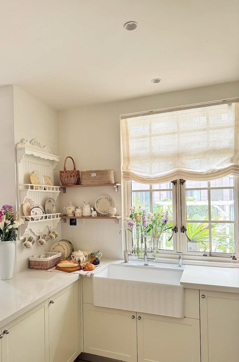 May include: A light and airy kitchen with white cabinets, a farmhouse sink, and a window with a linen blind. Decorative shelves display plates, mugs, and baskets. Fresh flowers in vases add colour to the space.