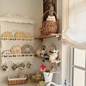 May include: A kitchen display with a white shelf holding decorative plates and mugs. A wicker basket contains towels, and a vase of pink flowers is on the counter. A floral painting hangs on the wall.