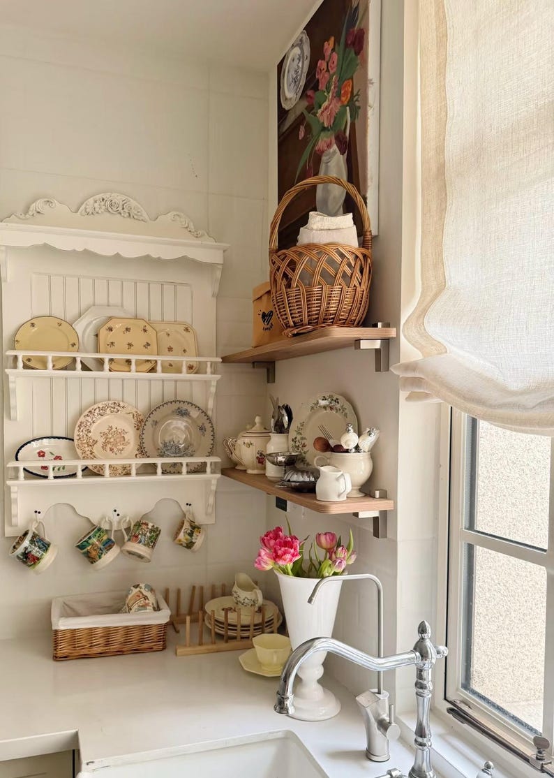 May include: A kitchen display with a white shelf holding decorative plates and mugs. A wicker basket contains towels, and a vase of pink flowers is on the counter. A floral painting hangs on the wall.