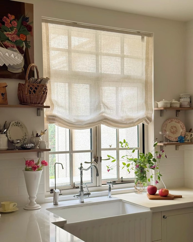May include: A kitchen interior featuring a white sink, window, and work surface. A woven basket, vase of flowers, and apples on a chopping board add colour. A beige window blind is drawn up, allowing natural light.