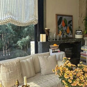May include: A cozy living room scene with a cream-colored sofa adorned with pillows. A large window with striped blinds overlooks a lush green landscape. A framed artwork and a vase of yellow flowers add decorative touches.