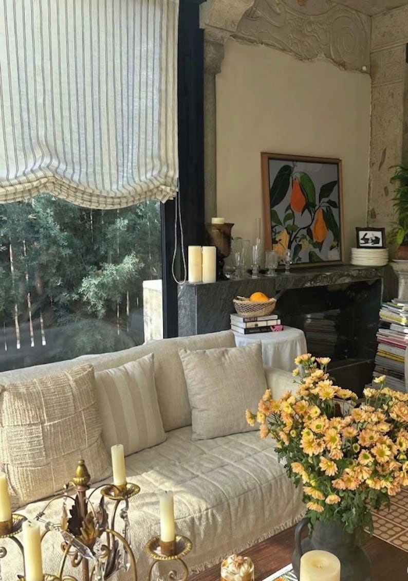 May include: A cozy living room scene with a cream-colored sofa adorned with pillows. A large window with striped blinds overlooks a lush green landscape. A framed artwork and a vase of yellow flowers add decorative touches.