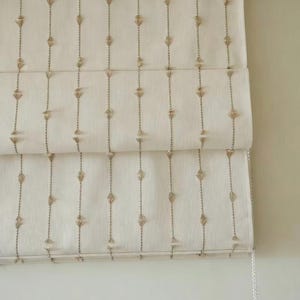 May include: Cream-colored Roman shade with vertical rows of embroidered diamond and dot patterns. The shade is partially rolled up, revealing its layered construction and a white pull cord.