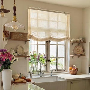 Peut inclure: Intérieur de cuisine lumineux avec une fenêtre ornée d'un store romain beige. Un évier blanc est devant la fenêtre, avec un bouquet de fleurs dans un vase. Un panier de fruits est posé sur le comptoir, et deux suspensions pendent du plafond.