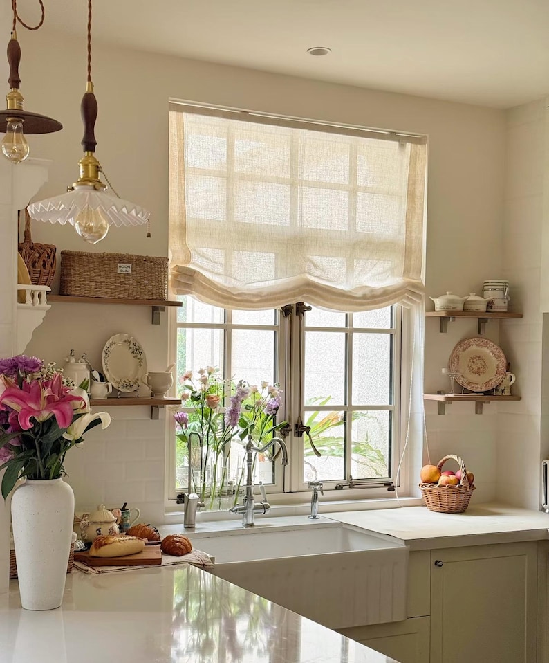 May include: A kitchen interior with a window covered by a beige Roman blind. A white sink is positioned in front of the window, with a vase of flowers. A basket of fruit rests on the counter, and two pendant lights hang from the ceiling.