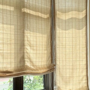 Puede incluir: Persianas romanas beige con un patrón de cuadrícula parcialmente desplegadas, revelando una ventana y la vista exterior. Las persianas están hechas de una tela de color claro y tienen una textura sutil. El marco de la ventana es marrón oscuro.