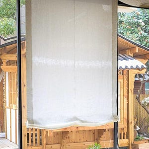 May include: A cream-colored, rectangular window shade made of a textured fabric. The shade is hanging in front of a window, with a wooden structure visible through the glass. The shade is pulled down, covering most of the window.