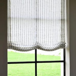 May include: A window with a white and gray striped Roman shade. The shade is partially drawn, revealing a green lawn outside. The window frame is black, and the walls are white.