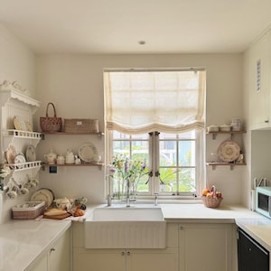 May include: A light and spacious kitchen featuring white cabinetry, work surfaces, and a farmhouse sink. Open shelves showcase decorative plates, teacups, and baskets. A window with a neutral blind overlooks a garden. A microwave and dishwasher are visible.