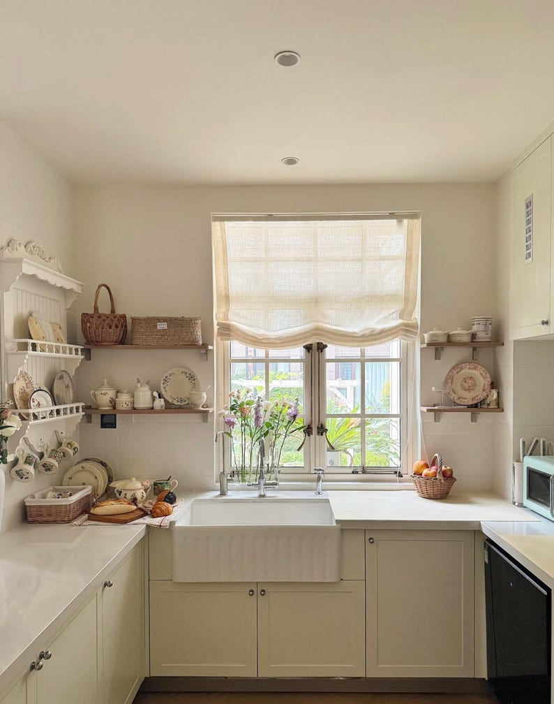 May include: A light and spacious kitchen featuring white cabinetry, work surfaces, and a farmhouse sink. Open shelves showcase decorative plates, teacups, and baskets. A window with a neutral blind overlooks a garden. A microwave and dishwasher are visible.