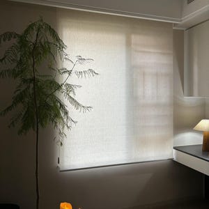 Pode incluir: Interior de um quarto com uma cortina de rolo em tons neutros em frente a uma janela. Uma pequena árvore num vaso branco está à esquerda, com duas velas acesas numa mesa pequena. Uma lâmpada é visível numa secretária próxima.