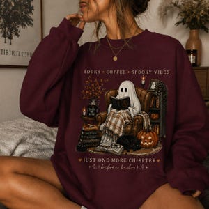 Könnte beinhalten: Ein bordeauxrotes Langarm-Sweatshirt mit einer Grafik eines Geistes, der in einem Sessel sitzt und ein Buch liest. Der Aufdruck enthält den Text BOOKS COFFEE SPOOKY VIBES und JUST ONE MORE CHAPTER before bed, umgeben von Kürbissen und Büchern.