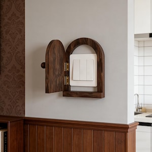 May include: A dark wood cabinet with an arched top and a small door, designed to conceal a white light switch panel. The cabinet is mounted on a light grey wall, with a glimpse of a kitchen in the background.