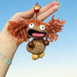May include: A handmade keychain depicting a character with a walnut shell head, orange yarn hair, and large white eyes. The character has a wide open mouth with a pink tongue and is adorned with brown spheres and yellow feet. A red apple slice tops the keychain.