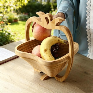 Op de afbeelding: Een houten fruitmand met een opvouwbaar ontwerp, met een banaan, twee appels en een tros druiven. De mand heeft een handvat en is gemaakt van lichtgekleurd hout. De mand staat op een houten tafel.