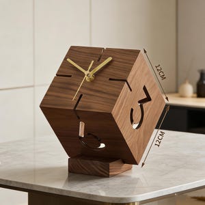 May include: A dark brown wooden cube clock with gold-coloured hands and cut-out numbers. Each side measures 12cm. The clock sits on a small, rectangular base. It is displayed on a marble surface.