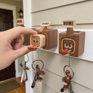 Puede incluir: Llaveros de madera con un diseño caprichoso. Cada soporte presenta una pequeña cara cuadrada con una expresión guiñadora y pequeños zapatos rojos. La palabra "HOME" se muestra en un pequeño letrero encima de cada soporte. Los llaveros cuelgan debajo.
