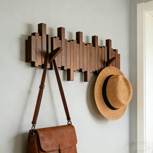 May include: A wooden coat rack fixed to a wall, featuring a distinctive design. The rack is constructed from vertical wooden slats of different heights and includes several hooks. A brown leather bag and a straw hat with a black band are suspended.