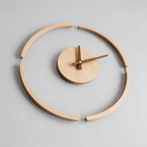 Könnte beinhalten: Eine minimalistische Wanduhr aus Holz mit hellem Holzgesicht und dunkelbraunen Zeigern. Das Design der Uhr umfasst einen kreisförmigen Rahmen aus gebogenen Holzstücken, die durch dünne Drähte verbunden sind und eine einzigartige, moderne Ästhetik schaffen. Der Durchmesser der Uhr beträgt ca. 30 cm.
