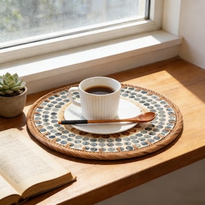 Puede incluir: Una taza de café blanca llena de líquido oscuro se encuentra sobre un platillo blanco, sobre un salvamanteles redondo decorativo. Una cuchara de madera con mango negro descansa sobre el platillo. También se ven una pequeña planta suculenta en maceta y un libro abierto.