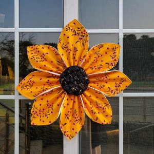 May include: A large fabric flower decoration featuring six bright yellow petals with a small red floral pattern and white trim. The center of the flower is a textured, rounded black fabric piece with a subtle gold-colored metallic thread pattern.
