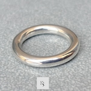 Peut inclure: Une bague argentée unie avec un design lisse et arrondi. La bague est circulaire et semble être en métal. La bague est présentée sur une surface grise.