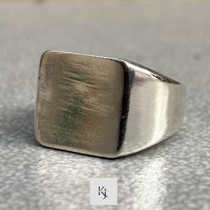 Op de afbeelding: Een zilveren zegelring met een vierkant vlak. De ring heeft een glad, gepolijst oppervlak en een dikke band. De ring is een klassiek sieraad, geschikt voor dagelijks gebruik. De ring wordt getoond op een grijze ondergrond.