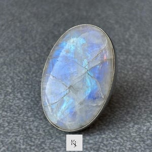 Puede incluir: Anillo de piedra lunar ovalado con una banda plateada. La piedra lunar tiene un color azul pálido y blanco con grietas internas y destellos iridiscentes. El anillo está sobre un fondo gris.