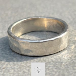 May include: A silver ring with a hammered texture. The ring has a flat band and a slightly rounded interior. The ring is shown on a grey surface. The ring is a jewellery item.