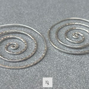 Könnte beinhalten: Ein Paar silberne Spiralohrringe. Jeder Ohrring hat eine gehämmerten Textur und ein Spiraldesign mit einem spitzen Ende zum Einsetzen. Die Ohrringe werden auf einer grauen Oberfläche präsentiert.