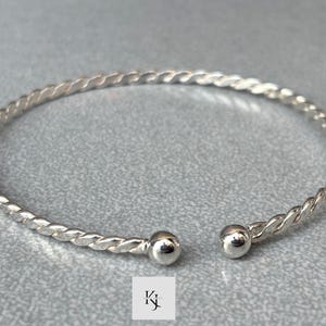 Könnte beinhalten: Ein silberfarbenes, offenes Armreif-Armband mit einem gedrehten Seil-Design. Das Armband hat an jedem offenen Ende eine glatte, abgerundete Kugel. Der Schmuck wird auf einer grauen Oberfläche präsentiert.