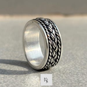 May include: A silver ring with a braided rope design around the band. The ring has a polished finish and a detailed texture, showcasing a classic and elegant style. The ring is a piece of jewelry.