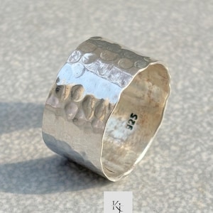 Può includere: Un anello largo in argento martellato. L'anello ha una superficie strutturata con una serie di piccole rientranze irregolari. L'interno dell'anello è liscio e lucido. Il numero "925" è stampato all'interno dell'anello.