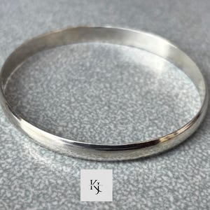 May include: A silver bangle bracelet. The round, solid bracelet is made of a shiny, light-coloured metal. The jewellery is displayed on a textured, grey surface. The bracelet is a simple, elegant design.