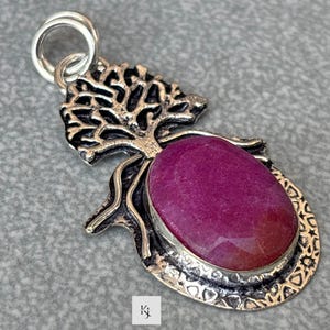 May include: Silver pendant with a large, oval-cut, deep pink gemstone. The pendant has a tree-like design at the top and a decorative border around the stone. A silver loop is at the top for a chain.