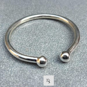 May include: A silver-colored open cuff bracelet with rounded ends and a smooth, polished finish. The bracelet has a simple, minimalist design, and the ends are capped with small, spherical accents. The bracelet is shown on a gray surface.