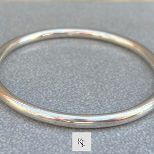 Puede incluir: Una pulsera de plata con un diseño liso y redondeado. La pulsera refleja la luz, creando reflejos y sombras. La joya es un accesorio sencillo y elegante.