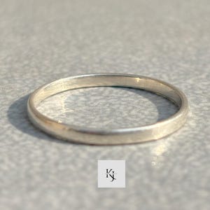 Può includere: Un anello a fascia d'argento. L'anello è una fascia sottile e semplice con una superficie liscia e lucida. L'anello è esposto su una superficie grigio chiaro. Un gioiello classico.