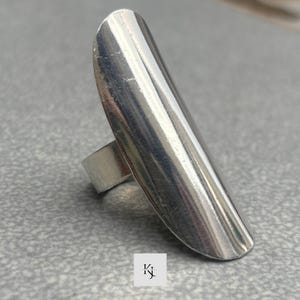 Pode incluir: Um anel prateado com um topo longo e oval. O anel tem uma superfície lisa e polida e uma banda simples. O anel está sobre um fundo cinzento.